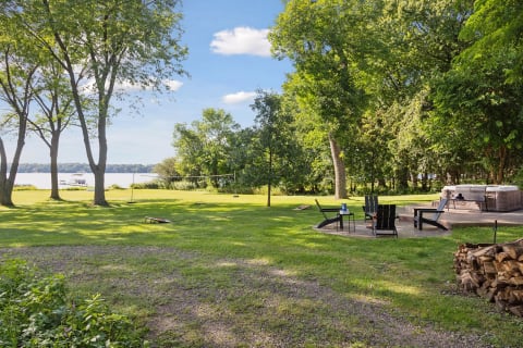 A peaceful outdoor area by a lake featuring green grass, seating, a hot tub, and trees under a blue sky.
