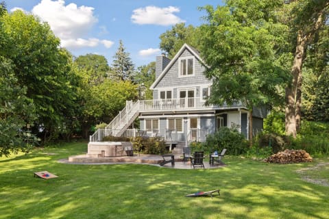 A two-story gray house with a wraparound balcony, landscaped yard, and outdoor seating area.