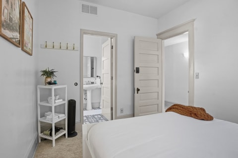 A cozy bedroom with a made bed, a throw blanket, a white shelving unit, and a view into a private bathroom.