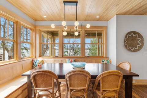 A modern dining room with large windows, wooden ceiling, and wicker chairs around a dark wooden table.