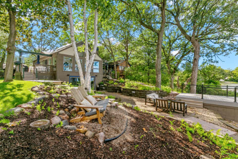 A cozy lakeside home surrounded by trees, featuring Adirondack chairs and a deck.