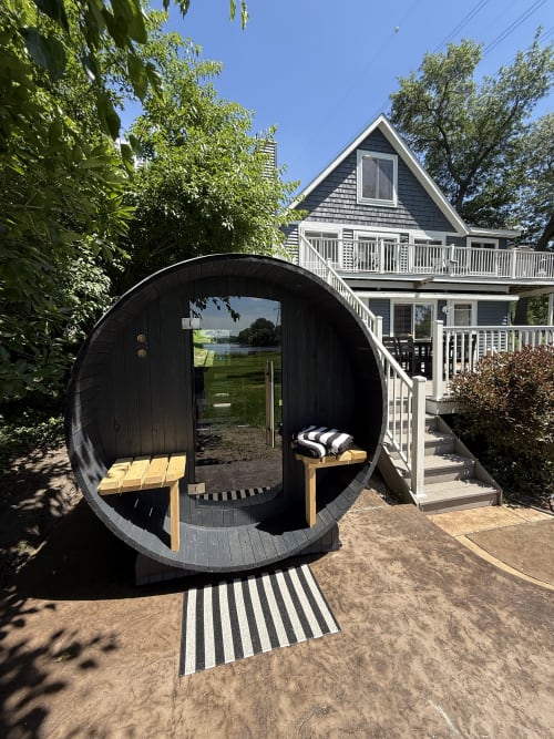 A unique barrel-shaped sauna awaits you in the garden, offering a cozy retreat surrounded by greenery and clear blue skies. Step inside to unwind in this distinctive outdoor wellness feature.