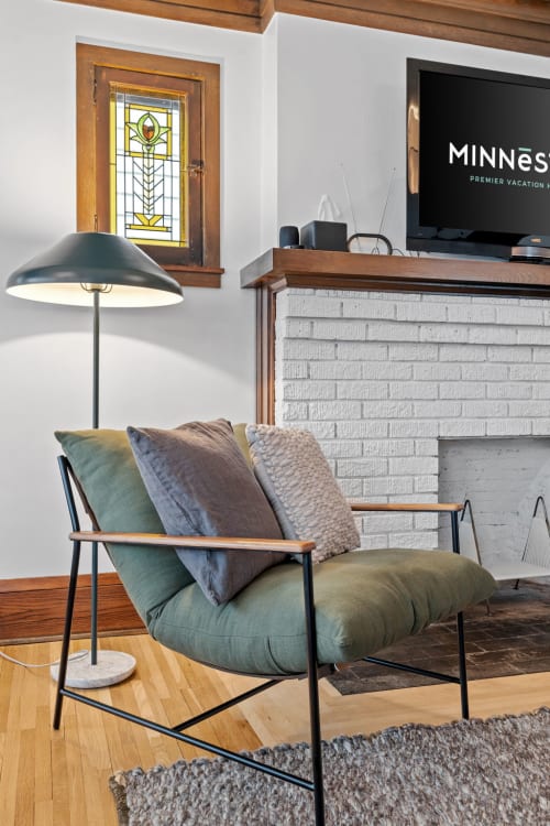 Sink into this plush green armchair by the fireplace, where warm wood meets modern comfort for your perfect evening unwind.