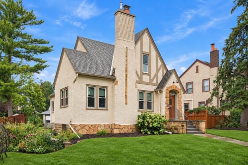 A charming Tudor-style home with distinctive stonework, surrounded by mature trees and lush greenery in a peaceful residential setting.